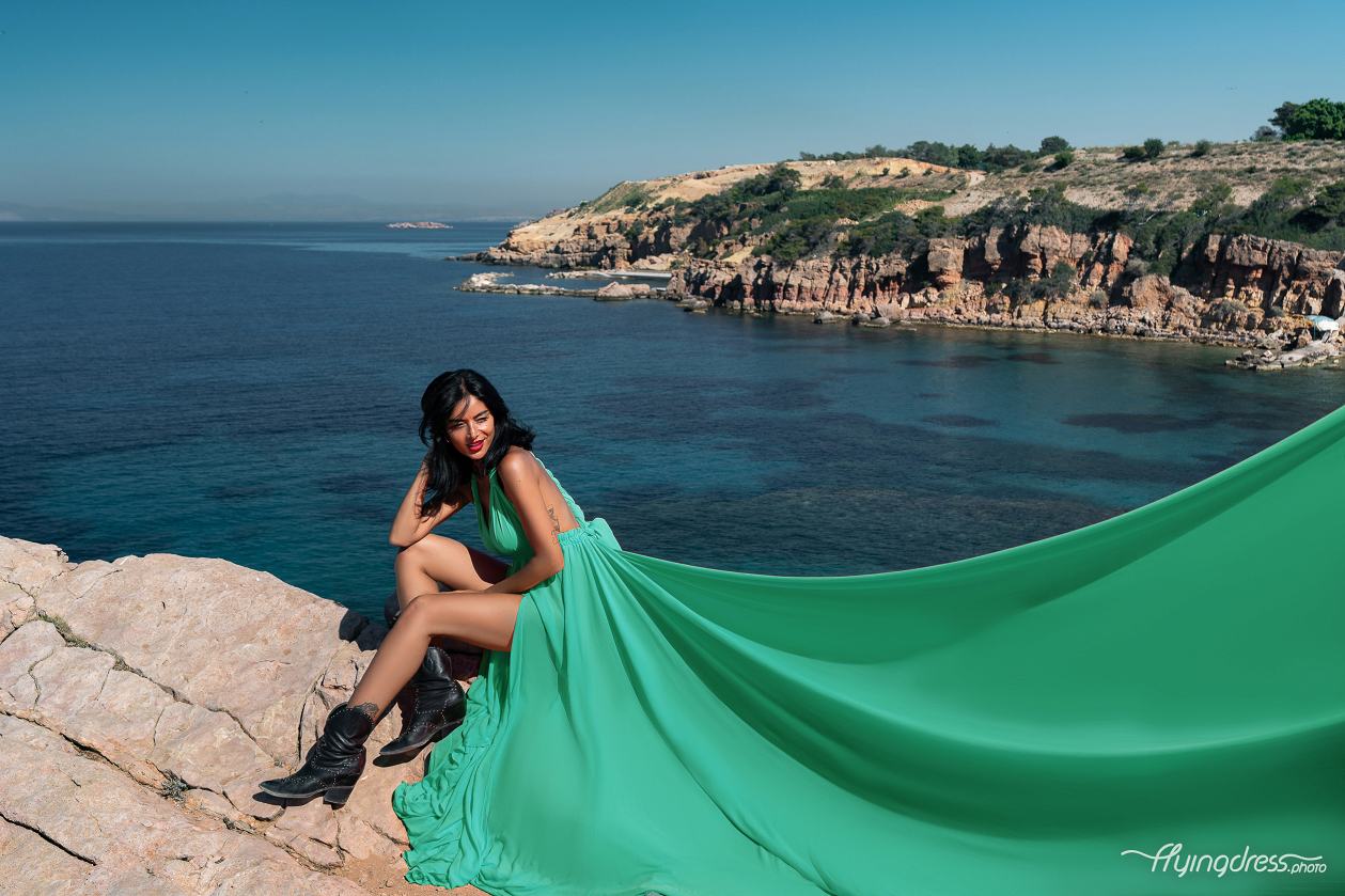 A woman in a flowing green dress sits on rocky cliffs by the sea in Vouliagmeni, Athens, with the coastline and clear blue waters stretching out behind her.